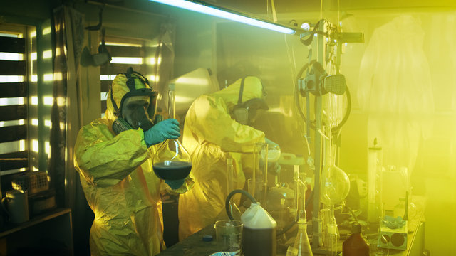 In the Underground Laboratory Two Clandestine Chemists Cook Drugs. They Wear Masks and Coveralls and Work with Beakers and Toxic Chemical Compounds.