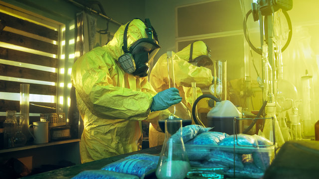 In the Underground Laboratory Two Clandestine Chemists Wearing Protective Coveralls and Masks Cook Drugs. They Work with Beakers, Distillation Glassware, Canisters and Hosepipe. True Crime Concept.