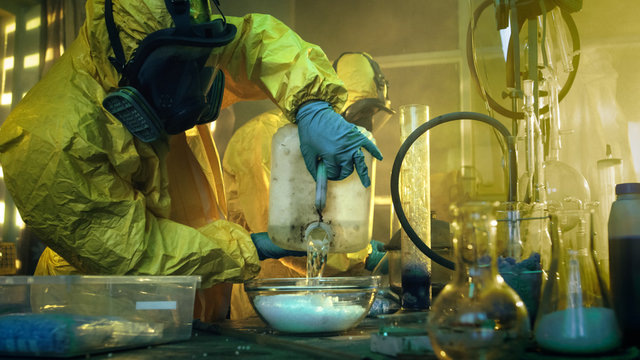In the Underground Drug Laboratory Two Clandestine Chemists Covered in Protective Coveralls and Gas Masks Mix Chemicals to Synthesise Drugs. They Work in the Abandoned Building.