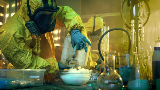 In the Underground Drug Laboratory Two Clandestine Chemists Covered in Protective Coveralls and Gas Masks Mix Chemicals to Synthesise Drugs. They Work in the Abandoned Building.