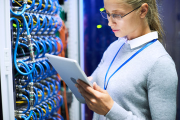 Portrait of female network engineer wearing glasses using digital tablet standing against server cabinets in data center