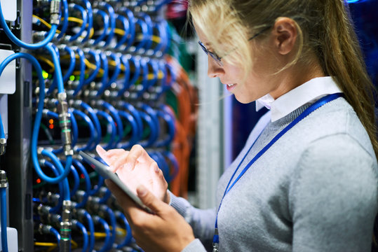 Side view portrait of young woman wearing glasses using digital tablet standing against server cabinets in data center