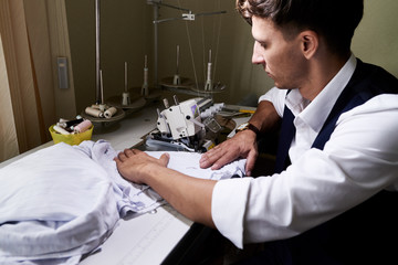 Side view portrait of handsome young tailor sawing clothes sitting at machine in atelier studio