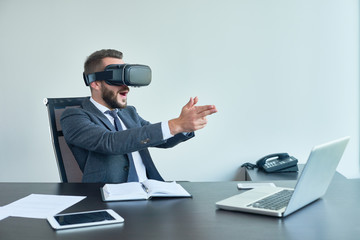 Portrait of handsome modern businessman using VR glasses playing shooter game sitting at desk in...