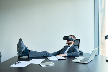 Portrait of handsome modern businessman wearing VR glasses relaxing at desk in office with feet on table
