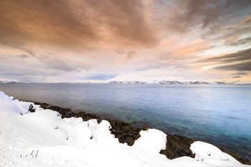 Sunset at the lakeside with rocks of a fjord during low tide in a snowy winter landscape.