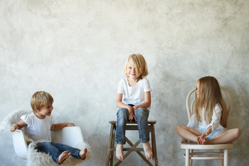Isolated shot of three cheerful children sitting on chairs in row along blank studio wall with copy space for your advertising content. Childhood, friendship, lesiure, fun, happiness and joy concept