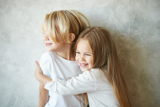 Two happy kids standing at blank grey wall and embracing. Adorable pretty little girl with long hair hugging tight cute blonde boy, showing her love and care. Brother and sister having fun at home