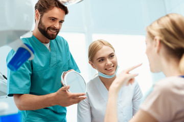 Obraz premium A woman is sitting in a dental chair. Before her stands a doctor and holds a mirror in her hands. Near the nurse