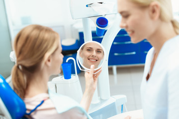 A woman is sitting in a dental chair and looking in the mirror. There is a dentist nearby