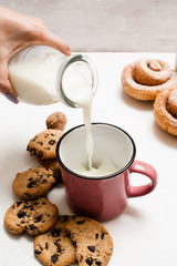Pastry organic breakfast with milk and scones, close up. Woman pouring fresh lacto drink into cup near wholegrain cookies and baked rolls laying on white table
