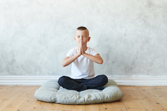 Isolated picture of happy little boy yogi sitting cross legged on mat on wooden floor in his bedroom, keeping palms pressed together and eyes closed, practising meditation after yoga exercise