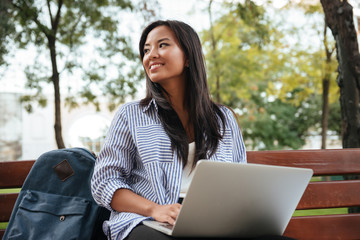 Portrait of young beautiful asian female student with laptop, sitting on wooden bench in park