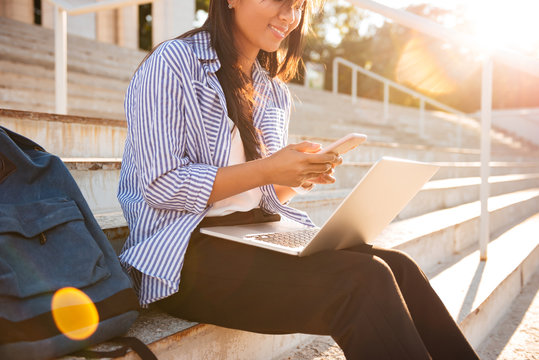 Photo of smiling asian female studen chatting on smartphone, while sitting on the cite stairs