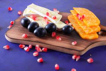 Camembert cheese, sliced cheese, orange cheese, marbled cheese, grains of pomegranate, dark blue grapes on a wooden board on a dark blue background in retro style