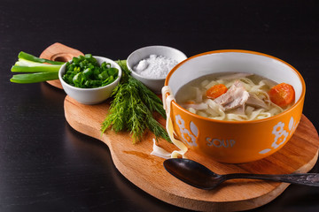 Homemade chicken noodle soup in a bowl on a wooden cutting board with salt shaker, chopped green onions and dill