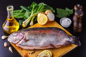 Fresh gutted trout on a wooden cutting board surrounded by ingredients for baking: olive oil, lemon, spring onions, garlic and spices. Top view