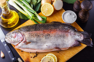 Fresh gutted trout on a wooden cutting board surrounded by ingredients for baking: olive oil, lemon, spring onions, garlic and spices. Top view
