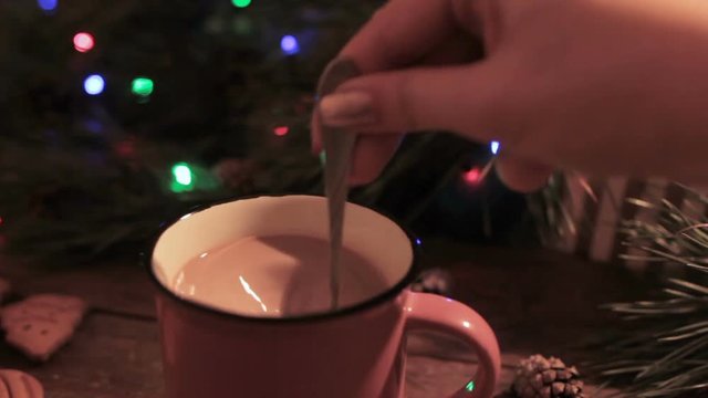 Delicious Christmas holiday with latte. Close up process of stirring up hot drink by unrecognizable woman on festive background of illuminated pine, gift box and gingerbread scone
