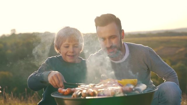 Father And Son Cook Sausages On Grill.