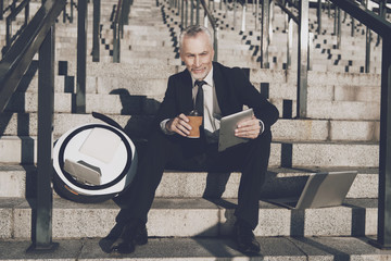 A respectable old man in a strict business suit sits on steps of office. He works on a tablet, next to his mono-wheel © Vadym Huzhva