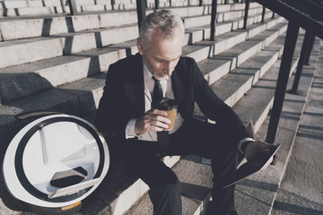 A respectable old man in a strict business suit sits on steps of office. He works on a tablet, next to his mono-wheel © Vadym Huzhva