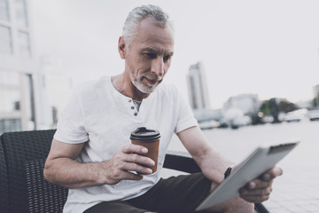 An old man with a beard sits on a sofa in the street. He is holding coffee and a tablet