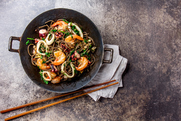 Buckwheat stir-fry noodles with seafood - shrimps, octopus, squid in cast iron asian wok with cooking chopstick. Top view, stone background - obrazy, fototapety, plakaty
