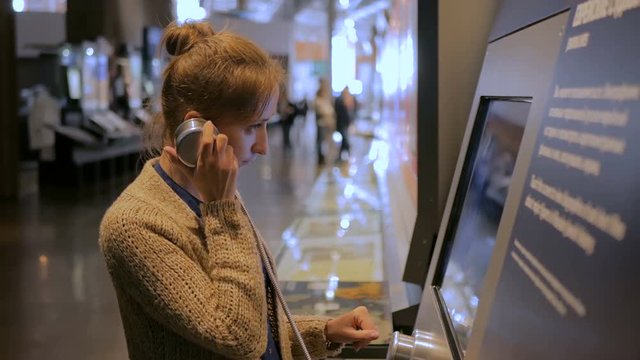 Woman touching interactive display, using headphones and listening audio guide at modern jewish history museum. Education and technology concept