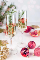 Sparkling cocktail with pomegranate and rosemary branch. New Year and Christmas Celebration. Two champagne glasses on light background. Red baubles, berries, garland, tree ornaments and gift boxes