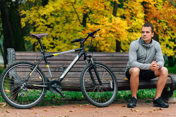 Obraz premium Young handsome athletic man standing with bicycle near lake in autumn park. Fall season background. Male cyclist outdoors.