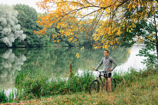 Young handsome athletic man standing with bicycle near lake in autumn park. Fall season background. Male cyclist outdoors.
