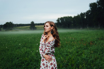 A beautiful girl in a colorful dress stands in the evening on a meadow against a background of fog.