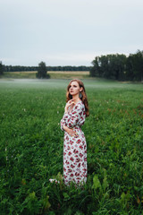 A beautiful girl in a colorful dress stands in the evening on a meadow against a background of fog.