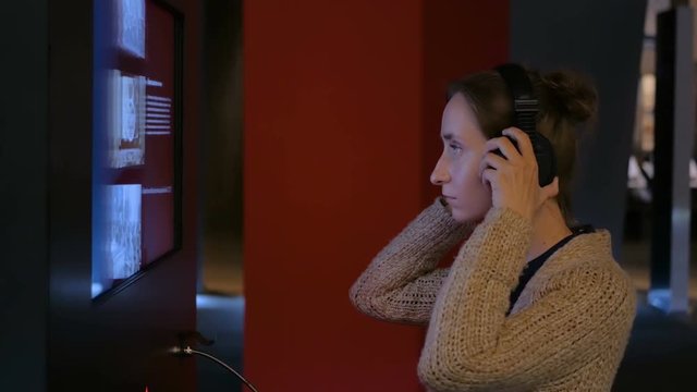 Woman touching interactive display, using headphones and listening audio guide at modern jewish history museum. Education and technology concept