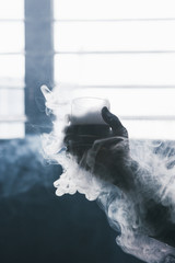 Creative alcoholic cold drink with white smoke. Glass with liquid in focus on foreground, bad habits addiction, unrecognizable male, dark atmosphere background