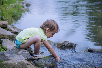 Adorable little child girl playing and having fun by leaves near mountain river on warm and sunny summer day. (Holiday, rest, happy childhood, games, nature concept)