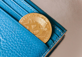 A blue leather wallet on a dark background with one gold and coin of bitcoin falling out of their...