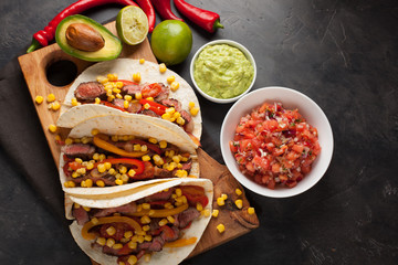 Three Mexican tacos with marbled beef, black Angus and vegetables on wooden Board on a dark stone background. Mexican dish with sauces guacamole and salsa in bowls. Top view
