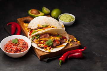 Three Mexican tacos with marbled beef, black Angus and vegetables on wooden Board on a dark stone background. Mexican dish with sauces guacamole and salsa in bowls. Top view