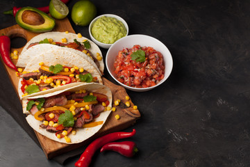 Three Mexican tacos with marbled beef, black Angus and vegetables on wooden Board on a dark stone background. Mexican dish with sauces guacamole and salsa in bowls. Top view with copy space