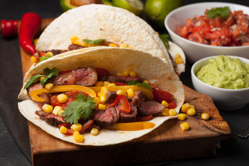 Three Mexican tacos with marbled beef, black Angus and vegetables on wooden Board on a dark stone background. Mexican dish with sauces guacamole and salsa in bowls