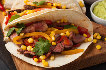 Three Mexican tacos with marbled beef, black Angus and vegetables on wooden Board on a dark stone background. Mexican dish with sauces guacamole and salsa in bowls