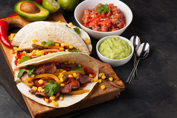 Three Mexican tacos with marbled beef, black Angus and vegetables on wooden Board on a dark stone background. Mexican dish with sauces guacamole and salsa in bowls. Top view