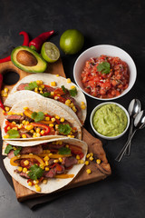 Three Mexican tacos with marbled beef, black Angus and vegetables on wooden Board on a dark stone background. Mexican dish with sauces guacamole and salsa in bowls. Top view