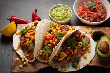 Three Mexican tacos with marbled beef, black Angus and vegetables on wooden Board on a dark stone background. Mexican dish with sauces guacamole and salsa in bowls