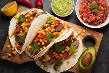 Three Mexican tacos with marbled beef, black Angus and vegetables on wooden Board on a dark stone background. Mexican dish with sauces guacamole and salsa in bowls