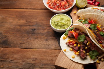 Three Mexican tacos with marbled beef, black Angus and vegetables on old rustic table. Mexican dish with sauces guacamole and salsa in bowls. Top view with copy space