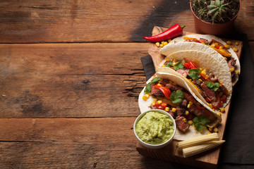 Three Mexican tacos with marbled beef, black Angus and vegetables on old rustic table. Mexican dish with sauces guacamole and salsa in bowls. Top view with copy space