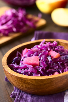 Braised red cabbage with apple in wooden bowl, with ingredients in the back, photographed with natural light (Selective Focus, Focus one third into the image)